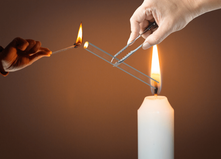 The glass tube is held against the candle flame with a lighted matchstick at its other end.